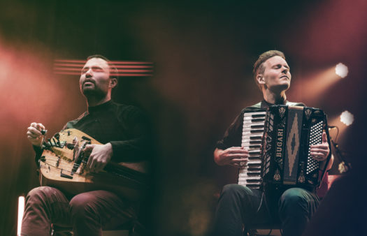 Members of music duo Symbio sit on chairs on-stage with their eyes closed and each playing their instrument, an accordion and a hurdy-gurdy.