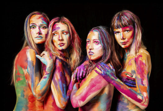 Upper body shot of four members of Tuuletar standing sideways with bare skin covered in colourful paint with hands on each others shoulders against a black background.