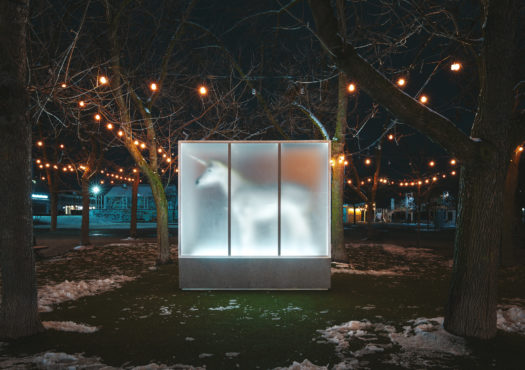 An outdoor art installation featuring a white unicorn inside of a translucent box lit from within with a silver metal frame and concrete foundation in a park at night.
