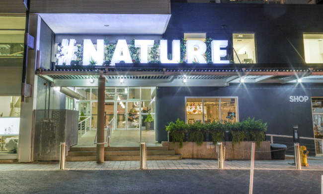 A large-scale art installation of #NATURE in three-dimensional glowing white block letters installed on the outside of a building at nighttime.