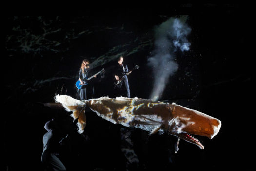 A darkly lit stage with a large whale puppet being held up by faintly seen people in black clothing. Steam rises from the whale's blowhole and two guitarists play upstage of it.
